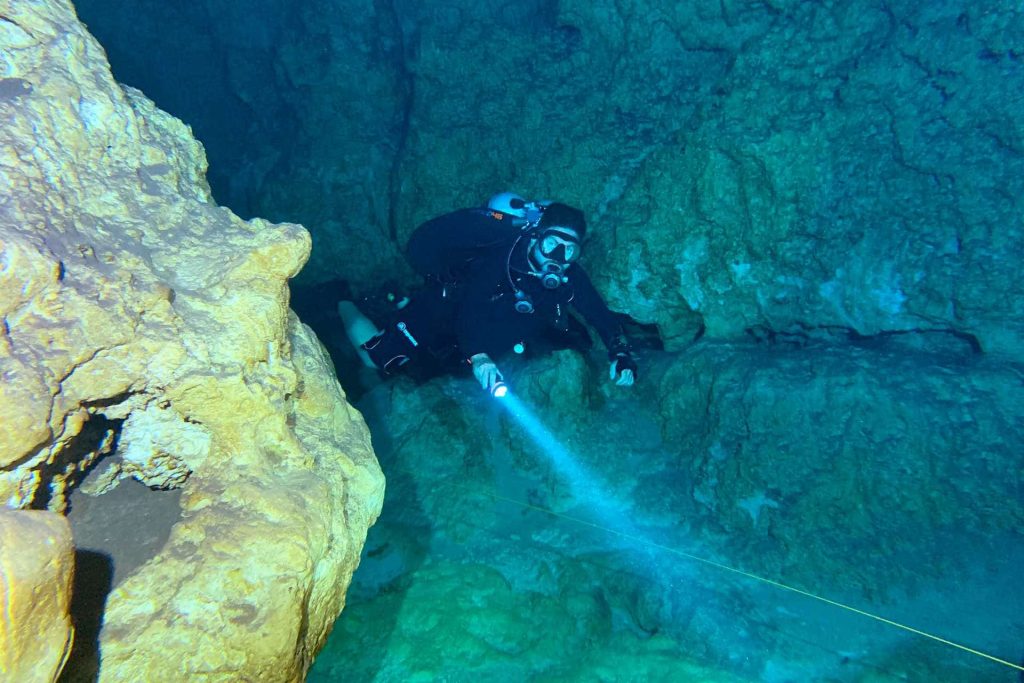 Cave diver wearing shorty and wing BCD holding lamp underwater