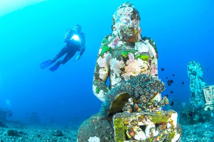 Scuba diver swimming besides statue underwater.